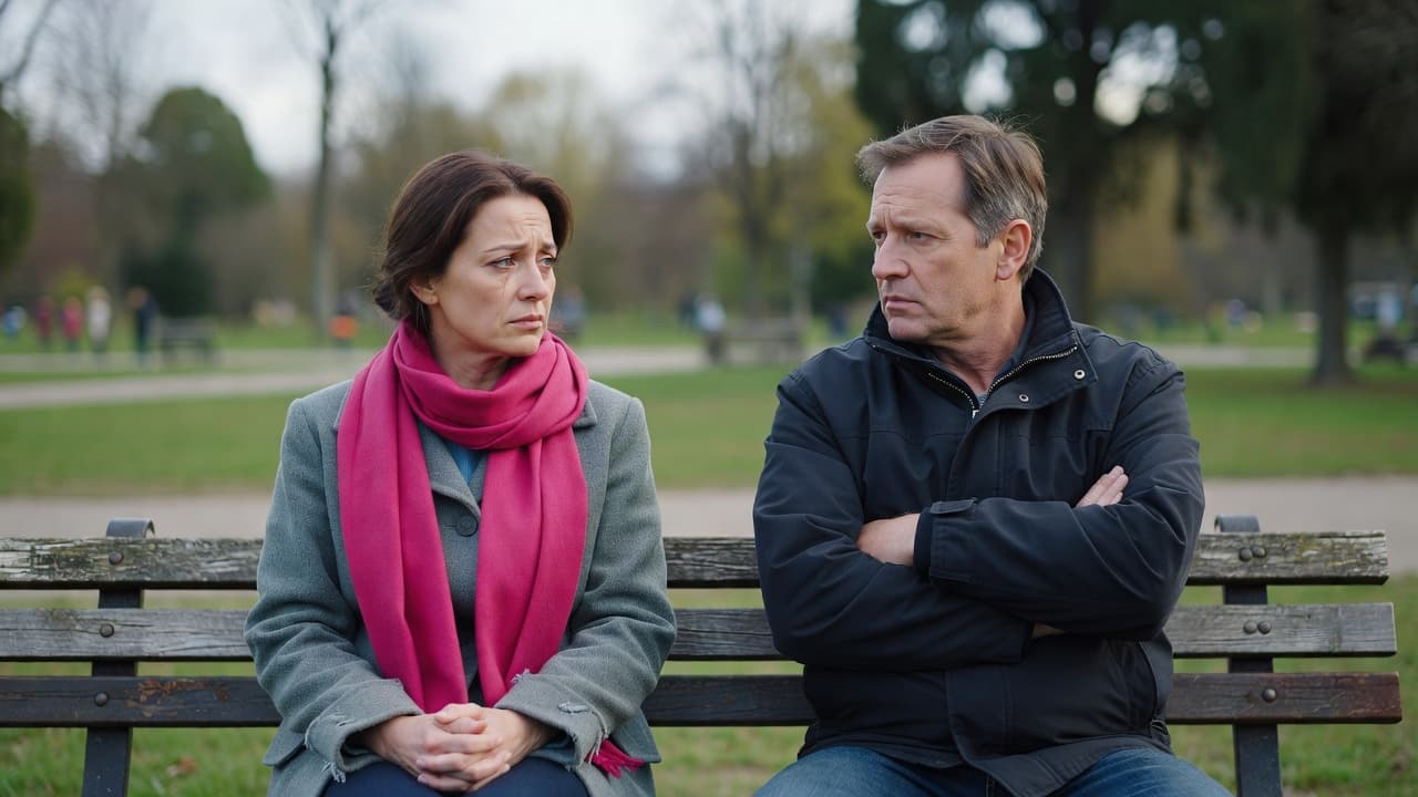 A man and woman sit on a wooden park bench engaged in a tense discussion. The woman wears a bright pink scarf as her expression conveys sadness during this serious moment.