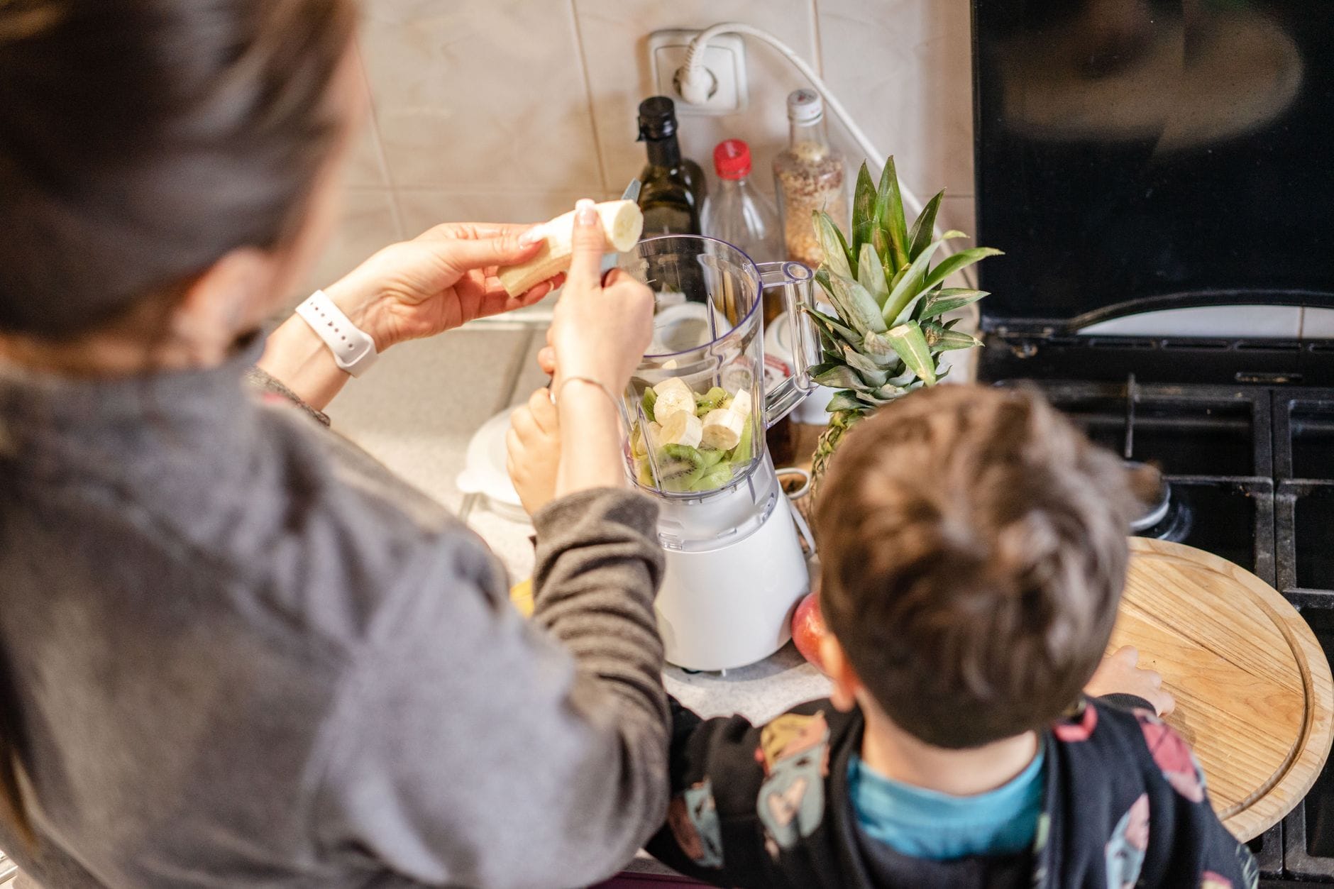 Mother with child making a healthy fruit smoothie in the kitchen, using bananas and pineapples.