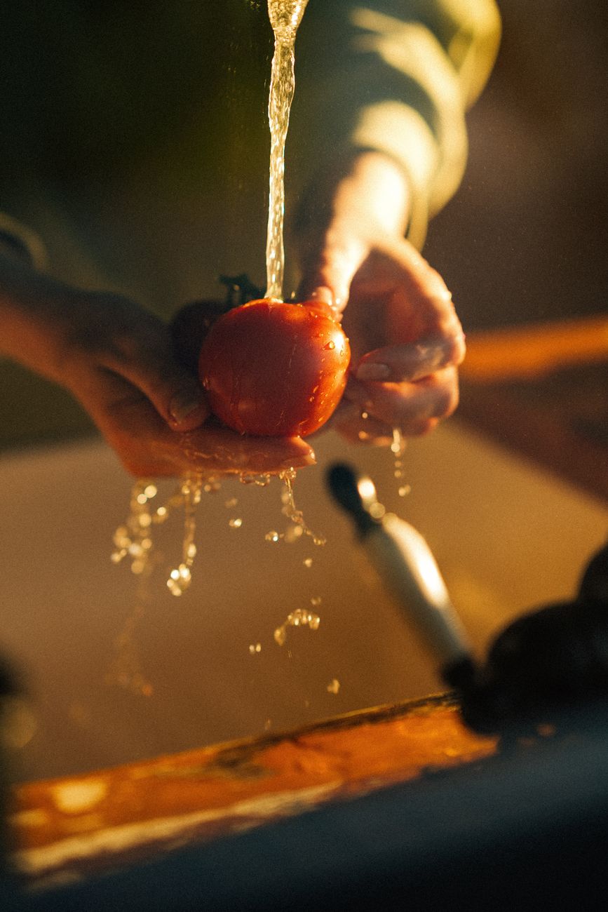 A person washing a fresh tomato under running water in a kitchen sink